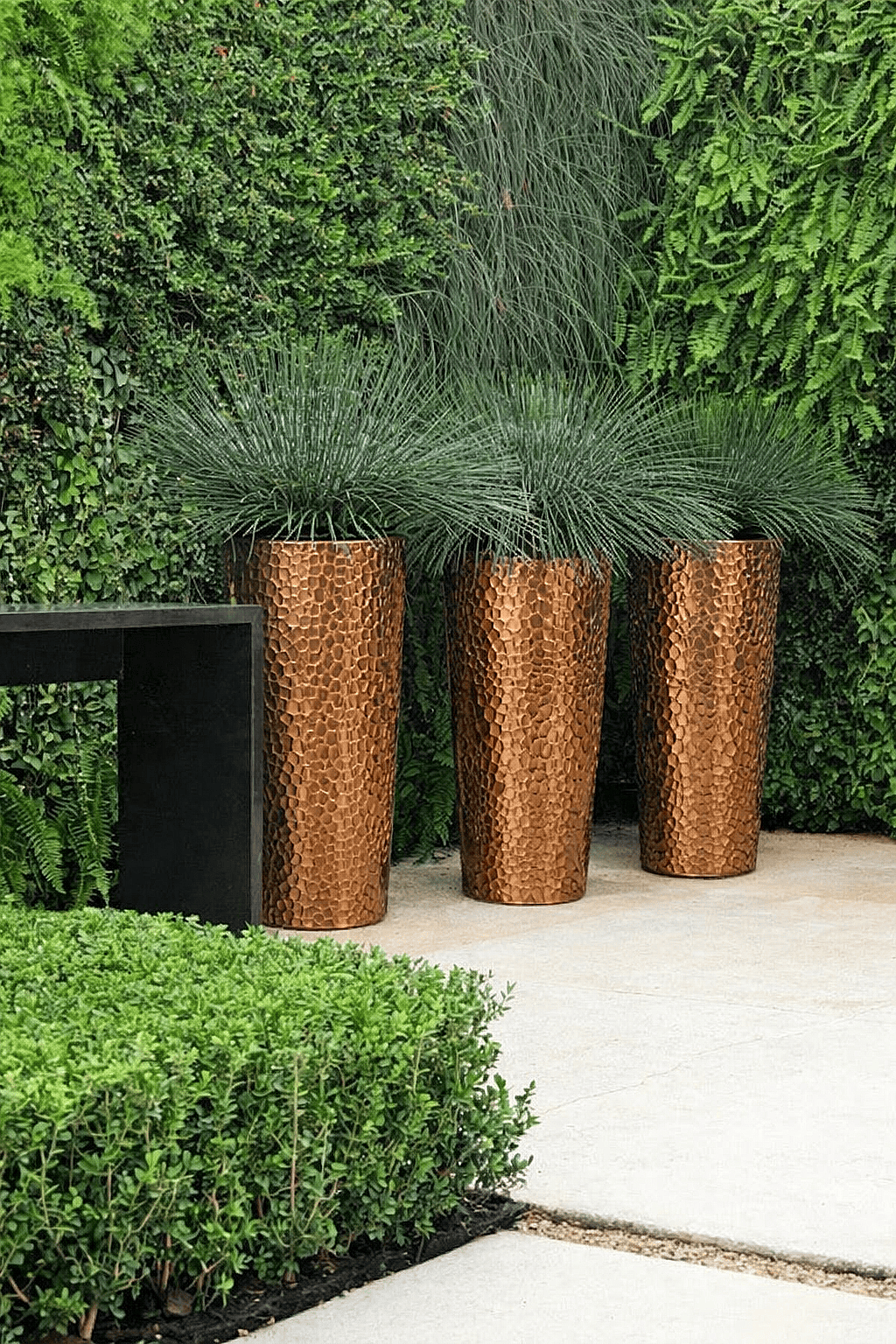 Three tall hammered metal planters with ornamental grasses arranged in a modern garden landscape