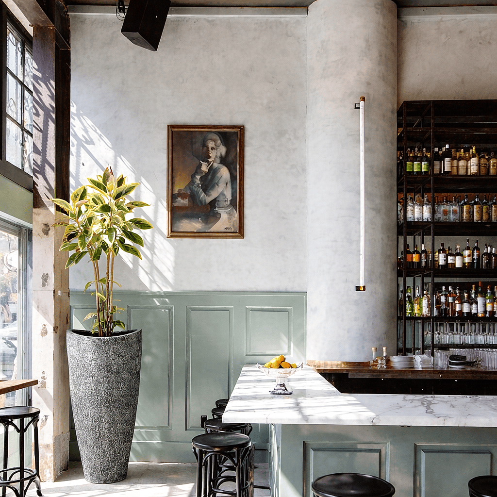 Tall tapered stone planter with indoor tropical plant placed beside a bar counter in a modern restaurant interior with large windows
